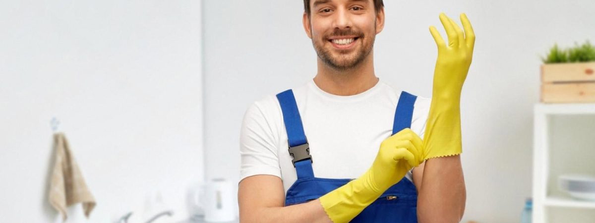 Smiling plumber in blue overalls putting on yellow gloves in a home kitchen